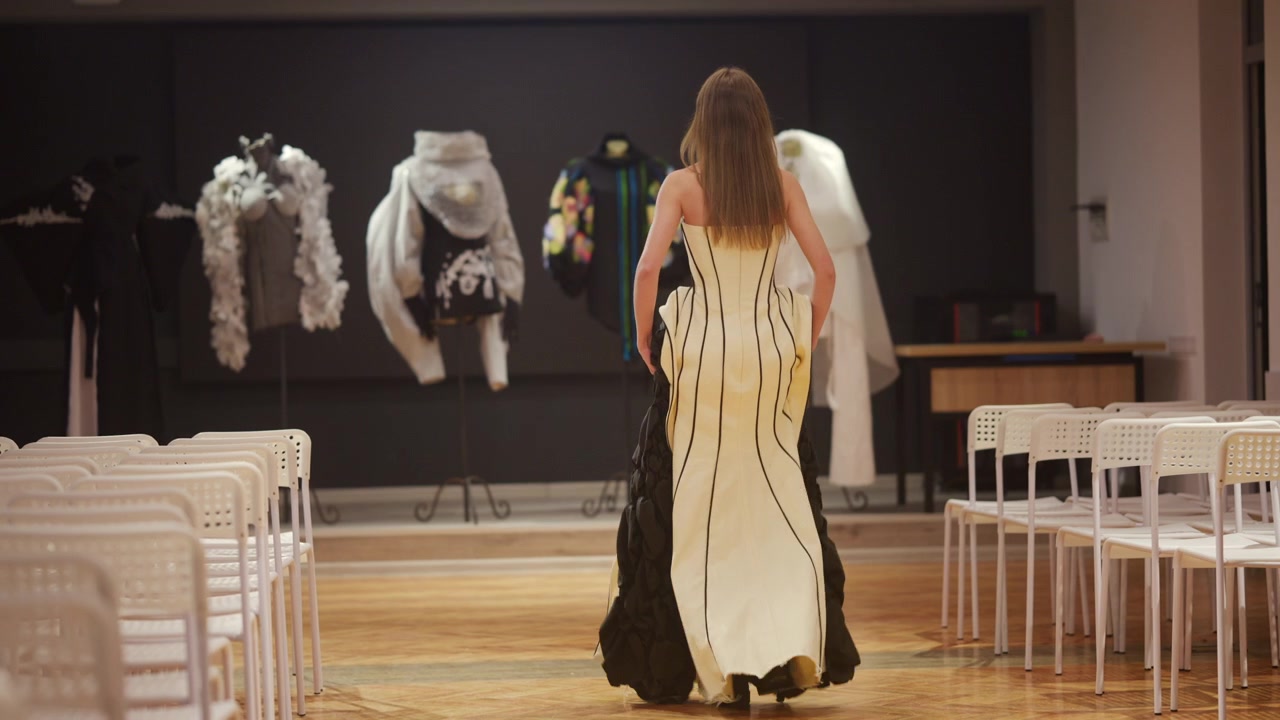 Vista posteriore di una modella che cammina indossando un abito strutturato stile corsetto con stecche nere verticali, presentato in una sfilata di moda minimalista al chiuso.