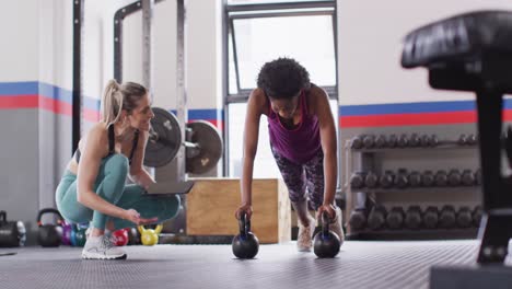 Video-of-diverse-female-fitness-trainer-instructing-woman-doing-push-ups-on-kettlebells-at-gym