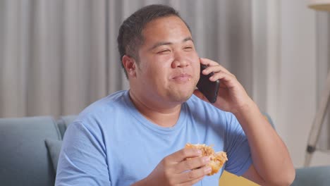 close up of a fat asian man talking on smartphone while eating fast food having hamburger on a sofa in the living room at home