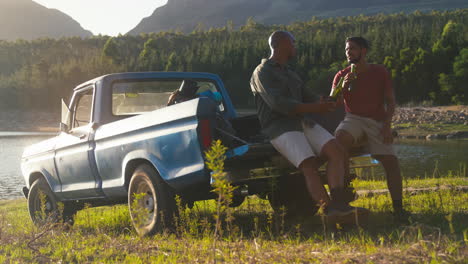 Two-Male-Friends-With-Backpacks-In-Pick-Up-Truck-On-Road-Trip-By-Lake-Drinking-Beer-And-Doing-Cheers