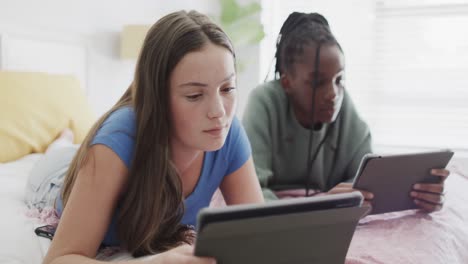 focused diverse teenage female friends lying on bed and using their tablets