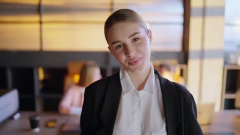 Portrait-of-a-happy-blonde-girl-in-a-black-business-uniform-posing-near-her-colleagues-from-the-table-in-a-sunny-office.-Confident-young-girl-businesswoman-posing-in-the-office