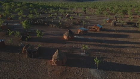 drone flies over a himba tribe camp in namibia during golden hour, revealing red earth, traditional huts, scattered trees, and distant mountains in stunning light.