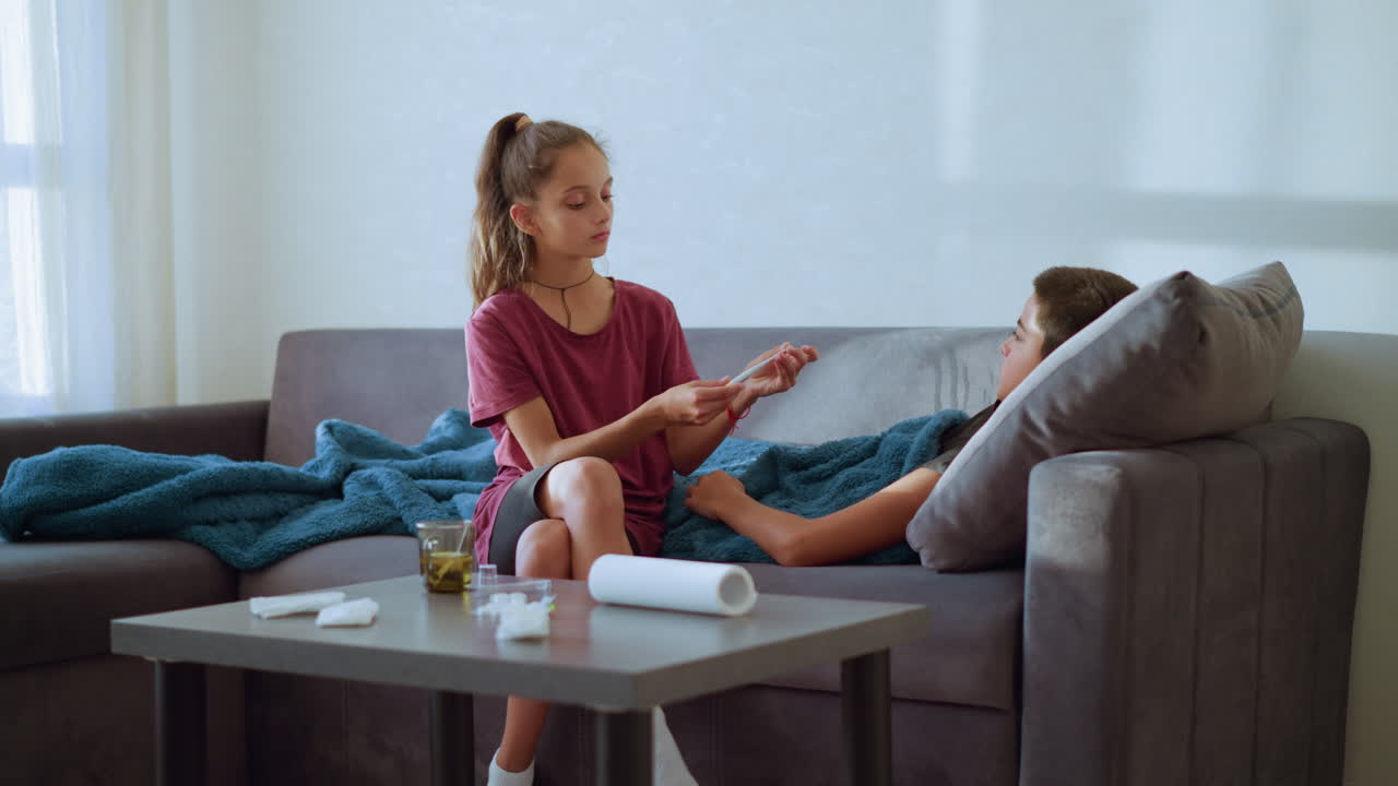 Girl checks thermometer used by her sick brother resting on couch while table holds tissue paper and glass cup in calm indoor scene filled with care concern