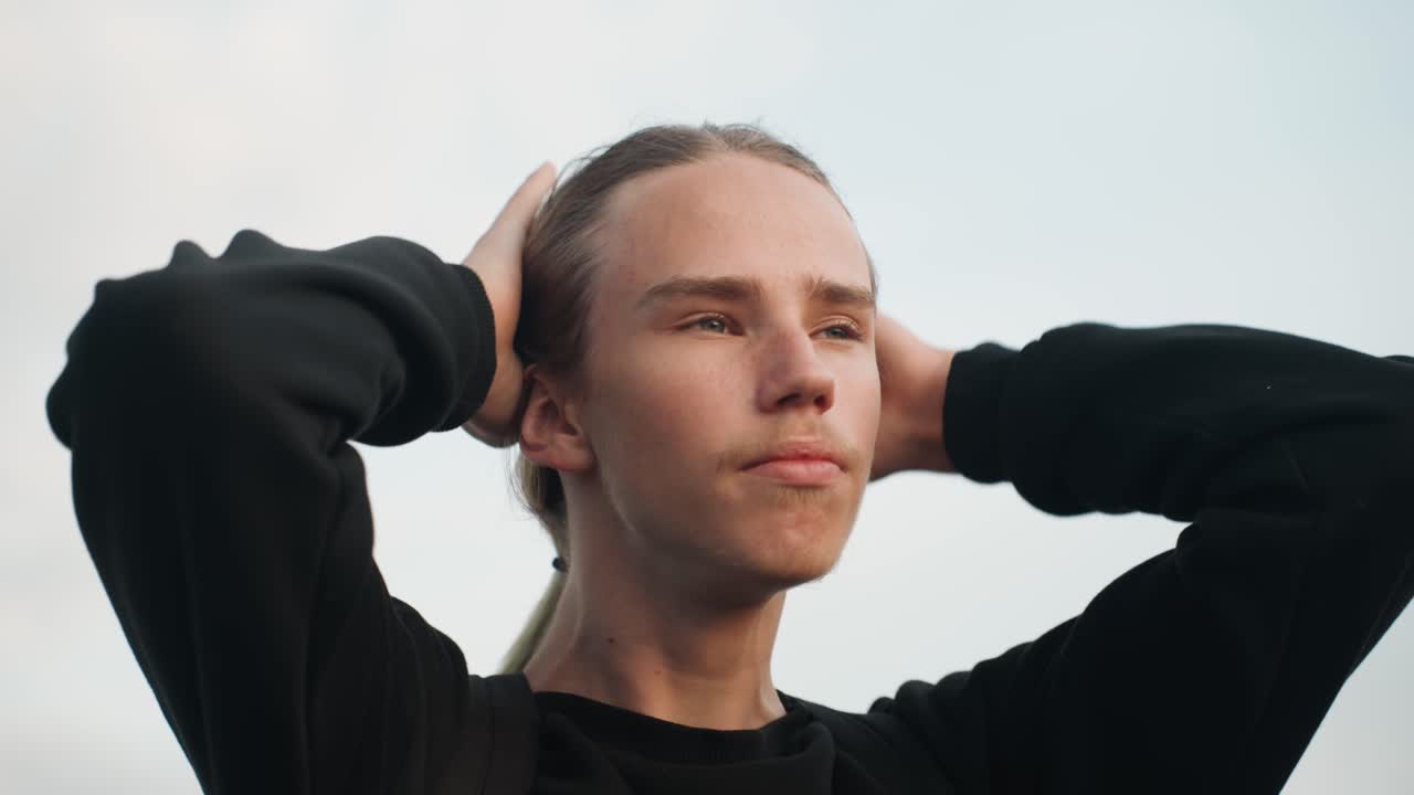 Giovane uomo caucasico che si liscia i capelli all'indietro con sguardo concentrato, mani che massaggiano il cuoio capelluto, postura eretta contro un cielo tenue, silhouette in maglione nero, primo piano di profilo che cattura una determinazione composta.