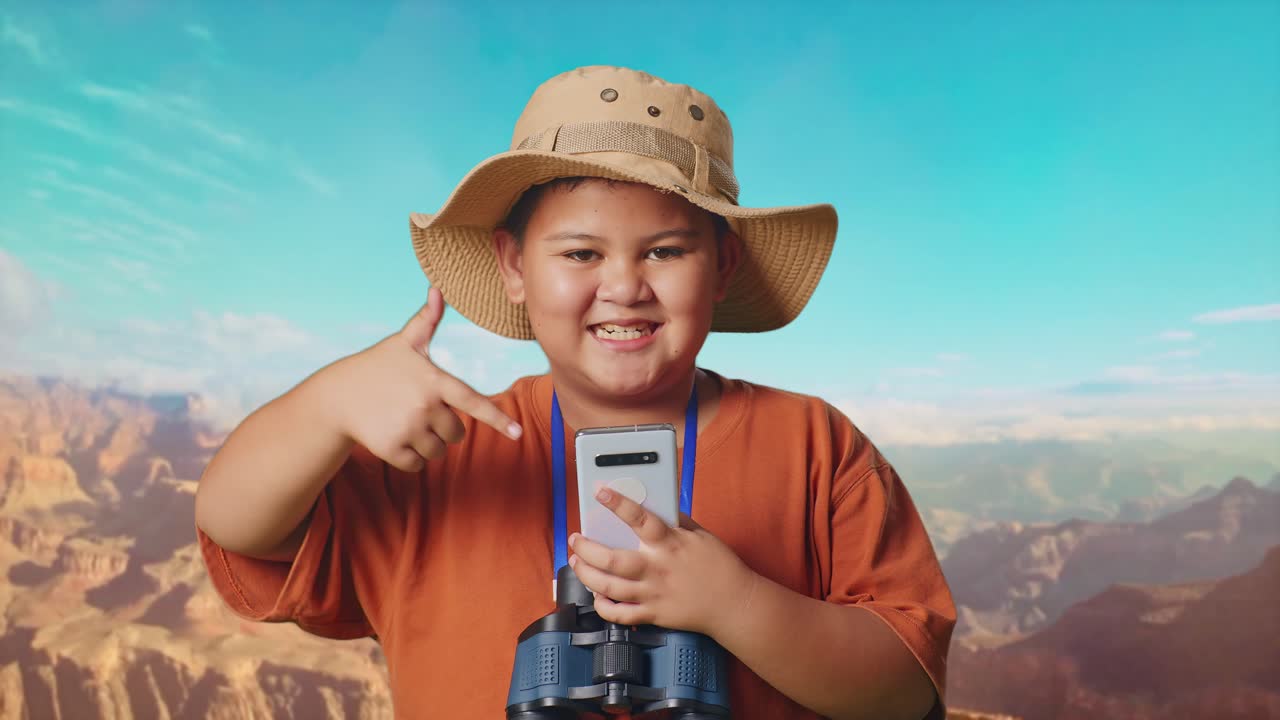 Asian Boy With A Hat And Binoculars Smiling And Pointing To Smartphone While Traveling At The Top Of Mountain. Boy Researcher Examines Something, Travel Tourism Adventure Concept, Close Up
