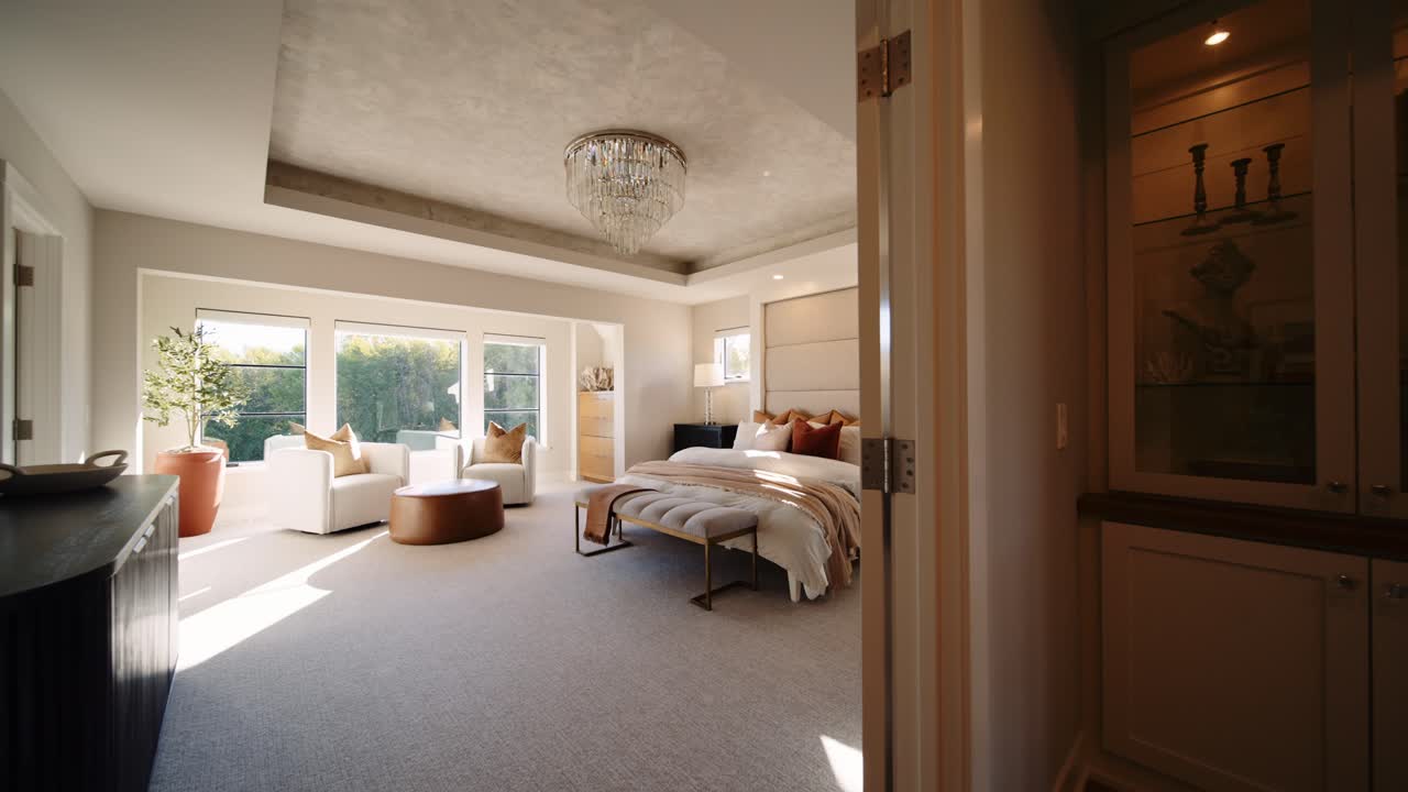 A luxurious bedroom with a large bed, cozy seating area, and chandelier, filled with natural light from multiple windows.