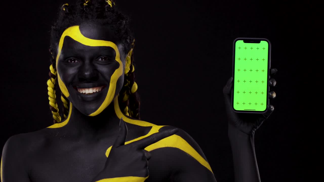 Face art. Woman with black and yellow body paint. Girl pointing finger at mobile phone screen with chroma key. Young african female with colorful bodypaint. An amazing afro american model with makeup.