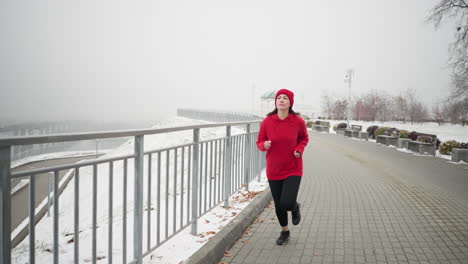 sportswoman in red beanie and jacket jogging near iron railing on snowy pathway surrounded by benches, foggy atmosphere, distant bridge, and serene winter park showcasing fitness in cold weather