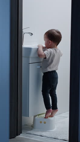 baby boy stands on toes to open water tap. kid is happy to watch the water flow. vertical video