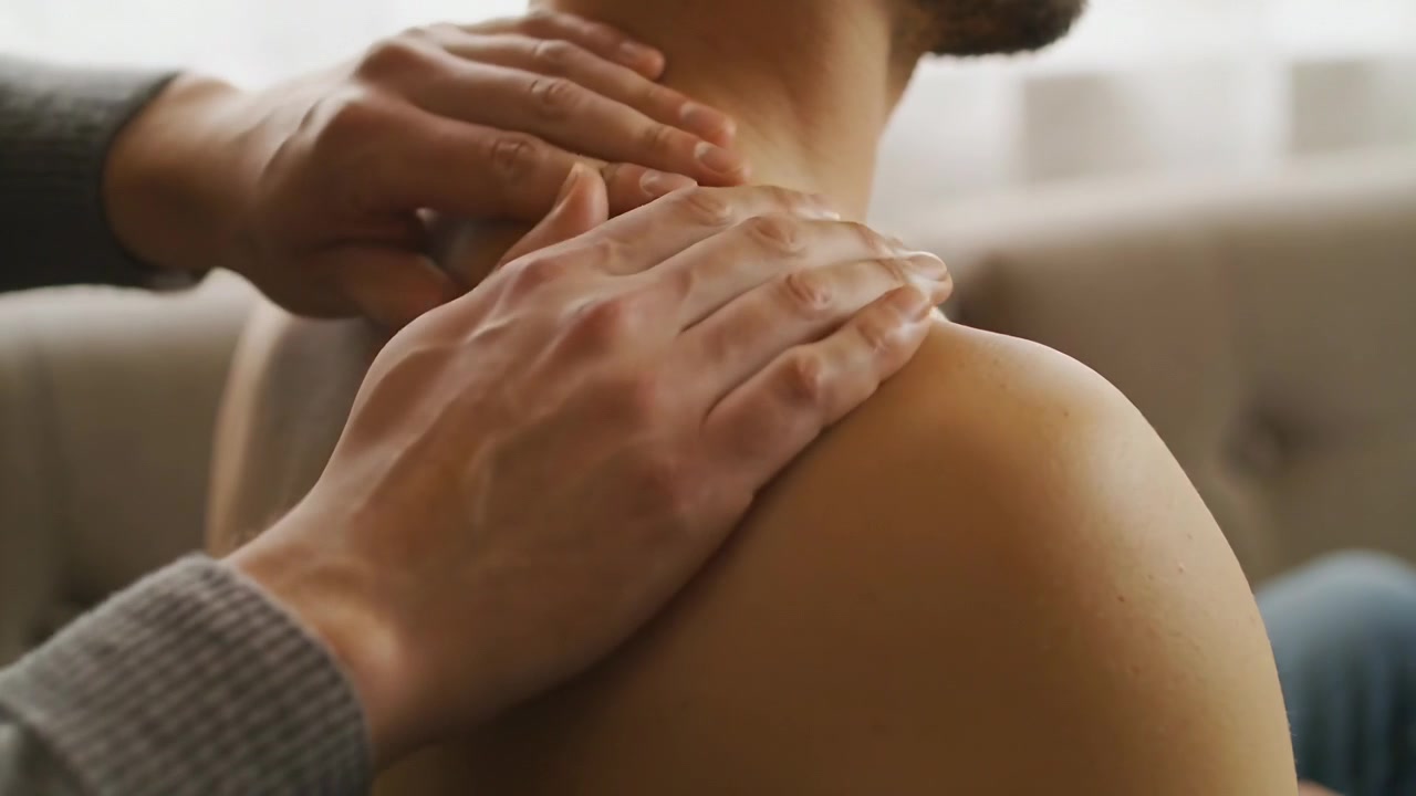 Mains vêtues d'un pull gris massant le cou et l'épaule d'un homme torse nu sur le canapé, soulageant la tension.