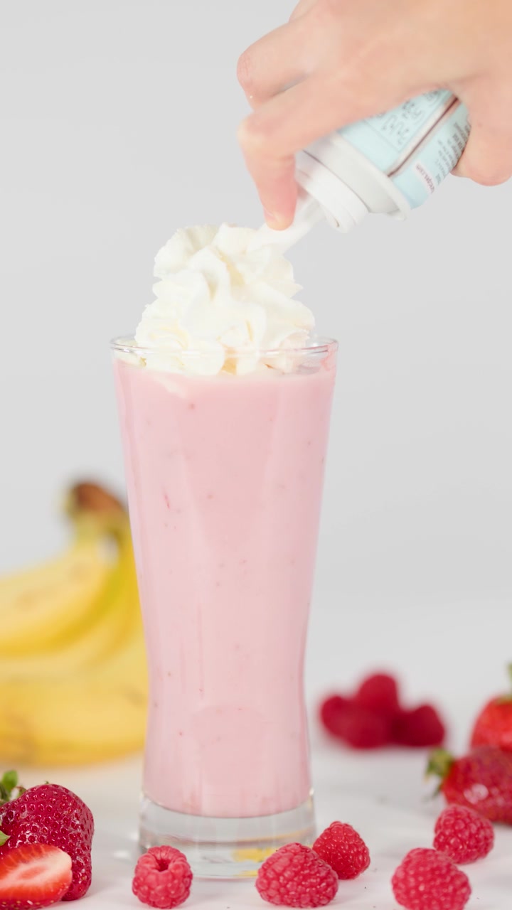 Una mano guarnisce un frullato di frutta rosa cremoso con panna montata, circondato da frutta fresca, sotto una luce brillante
