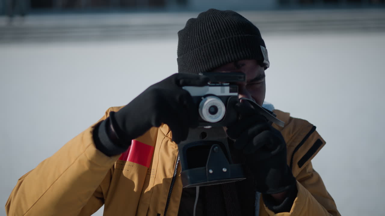 side view of black man peering through camera viewfinder and adjusting focus dial while reviewing shot on LCD display against snowy urban scene with blurred trees and parked cars under winter sun