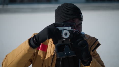 side view of black man peering through camera viewfinder and adjusting focus dial while reviewing shot on lcd display against snowy urban scene with blurred trees and parked cars under winter sun
