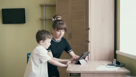 El-Seguimiento-De-Una-Joven-Y-Bonita-Madre-Le-Muestra-A-Su-Hijo-Cómo-Hacer-La-Tarea-Usando-Una-Tableta