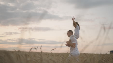 Daddy-carries-on-his-shoulders-his-beloved-little-healthy-daughter-in-sun.-In-slow-motion-the-daughter-walks-with-her-father-on-the-field-and-free-and-happy-waves-her-hands-up.-walking-in-field.