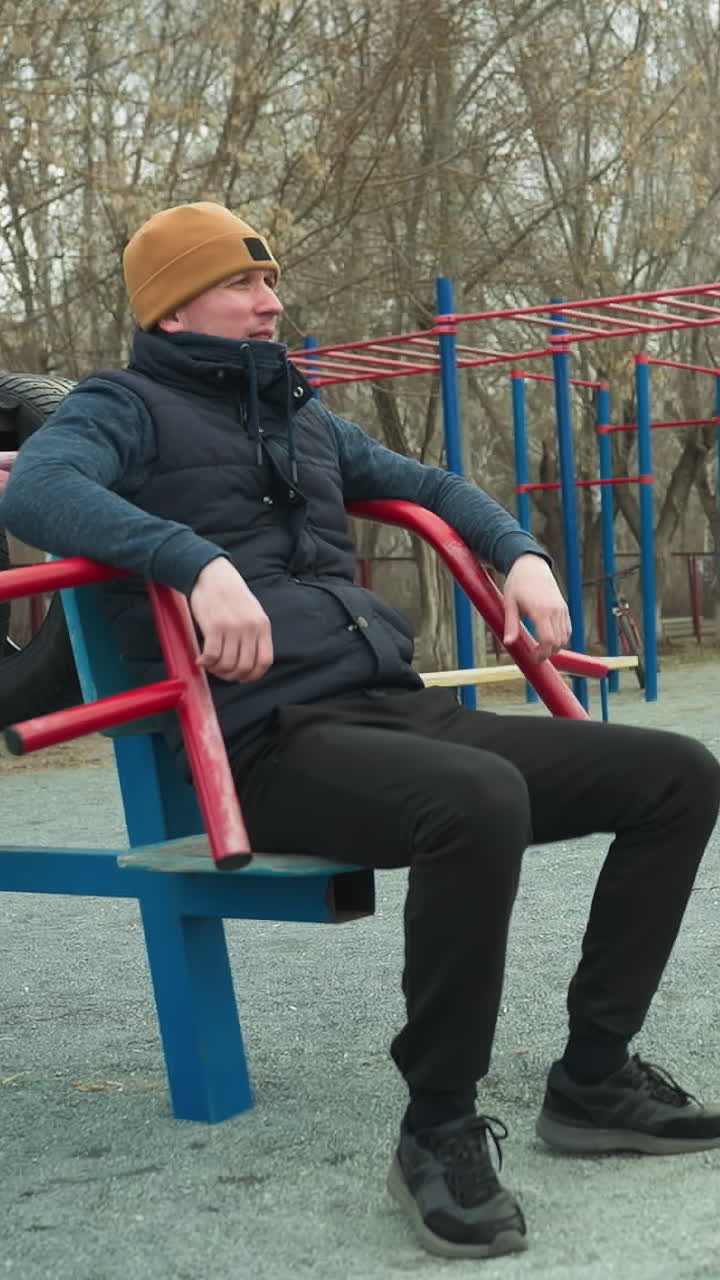 A coach is seated on workout equipment, casually shaking his leg, in the background, two boys are happily playing football and having a conversation, as one adjust backward