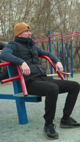 a coach is seated on workout equipment, casually shaking his leg, in the background, two boys are happily playing football and having a conversation, as one adjust backward