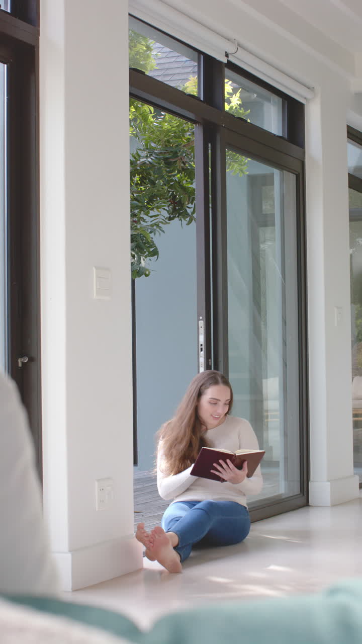 Vertical video of caucasian woman reading book in sunny living room, slow motion