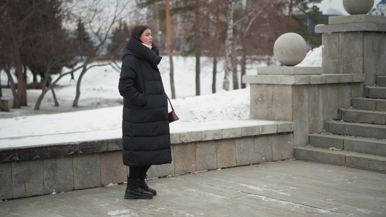 Donna con un lungo cappotto nero si trova all'aperto su un terreno coperto di neve vicino a gradini di pietra e sfere, guardandosi intorno. La donna indossa stivali con le mani parzialmente visibili sotto la patta della giacca. Sullo sfondo