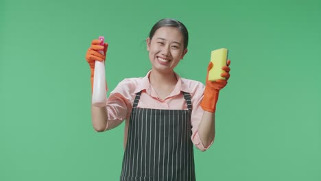 una ama de casa asiática sonriente con un delantal que muestra un spray y una esponja de cocina mientras está de pie en el estudio de pantalla verde