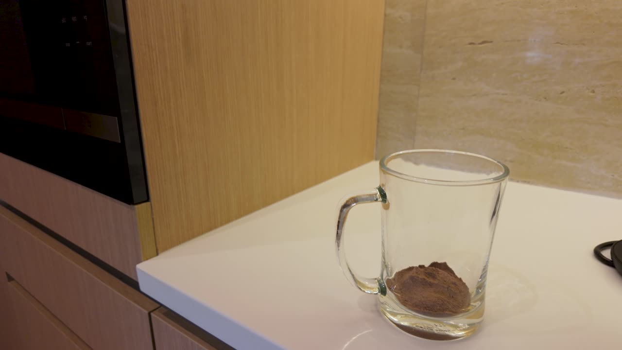 Making instant hot chocolate. A clear mug with cocoa powder beside a boiling kettle, warmly lit in a modern kitchen setting, evokes cozy anticipation