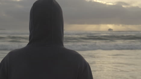 Rear-view-of-young-adult-hood-male-watching-boats-across-sunset-ocean-horizon