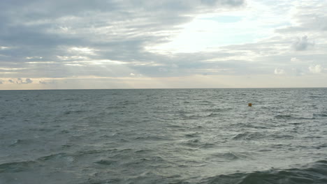 Boya-Amarilla-En-Un-Océano-Agitado-Con-Un-Paisaje-Nublado-Al-Atardecer-En-La-Distancia,-Avance-Aéreo