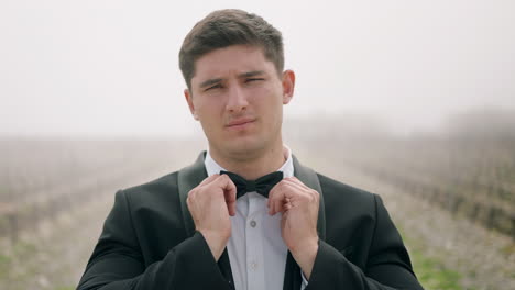 man in tuxedo adjusting bow tie in a foggy vineyard