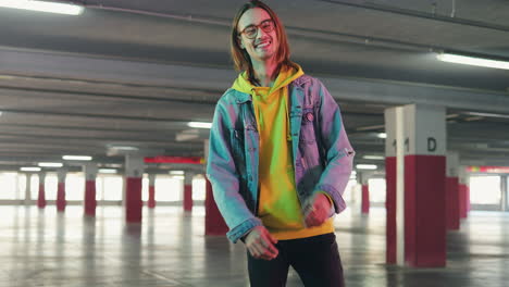 Stylish-young-male-hipster-in-glasses-standing-in-Aempty-parking,-looking-at-the-camera-and-crossing-his-arms