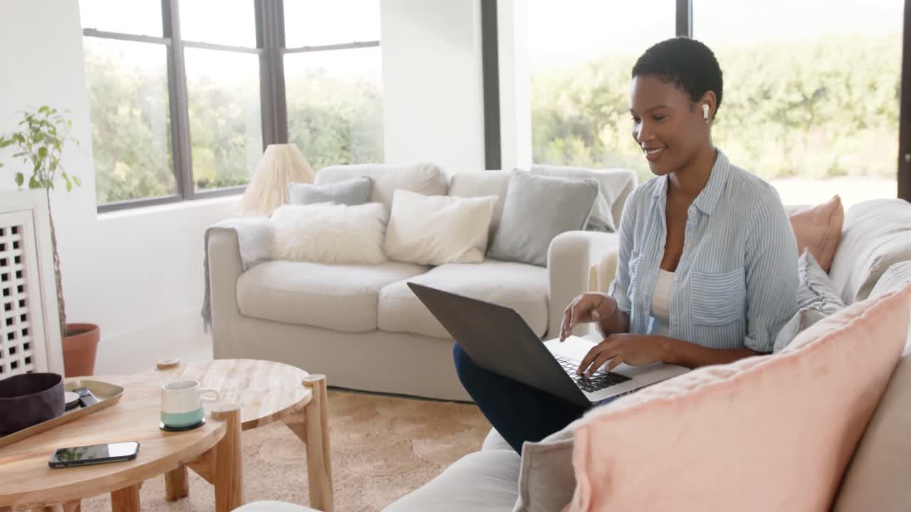 Happy african american woman sitting on sofa wearing earphones and using laptop, slow motion
