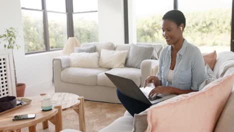 Happy-african-american-woman-sitting-on-sofa-wearing-earphones-and-using-laptop,-slow-motion