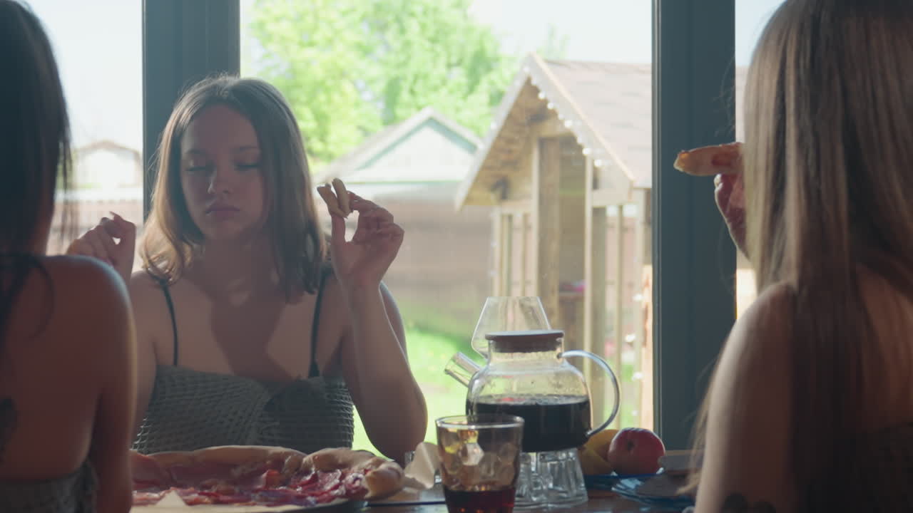 Back view of friends seated and eating while one cheerful lady in front enjoys herself, gesturing expressively and savoring a slice of pizza during a lively and relaxed mealtime in a sunny setting