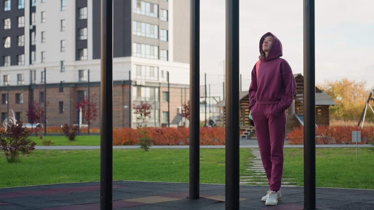 Training partner strides towards iron bar entrance pausing to gaze up at tall structure in urban outdoor gym park surrounded by autumn foliage city backdrop capturing dynamic fitness moment