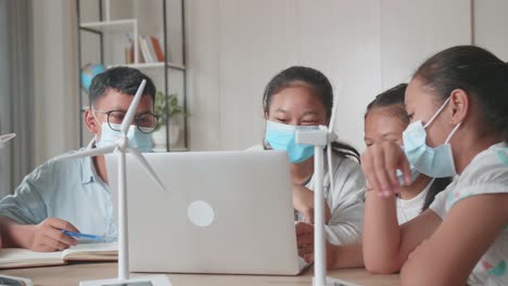 young asian children are wearing facemasks and work as a team using laptop computers to program wind turbines. classroom with kids learning about eco-friendly forms of renewable energy