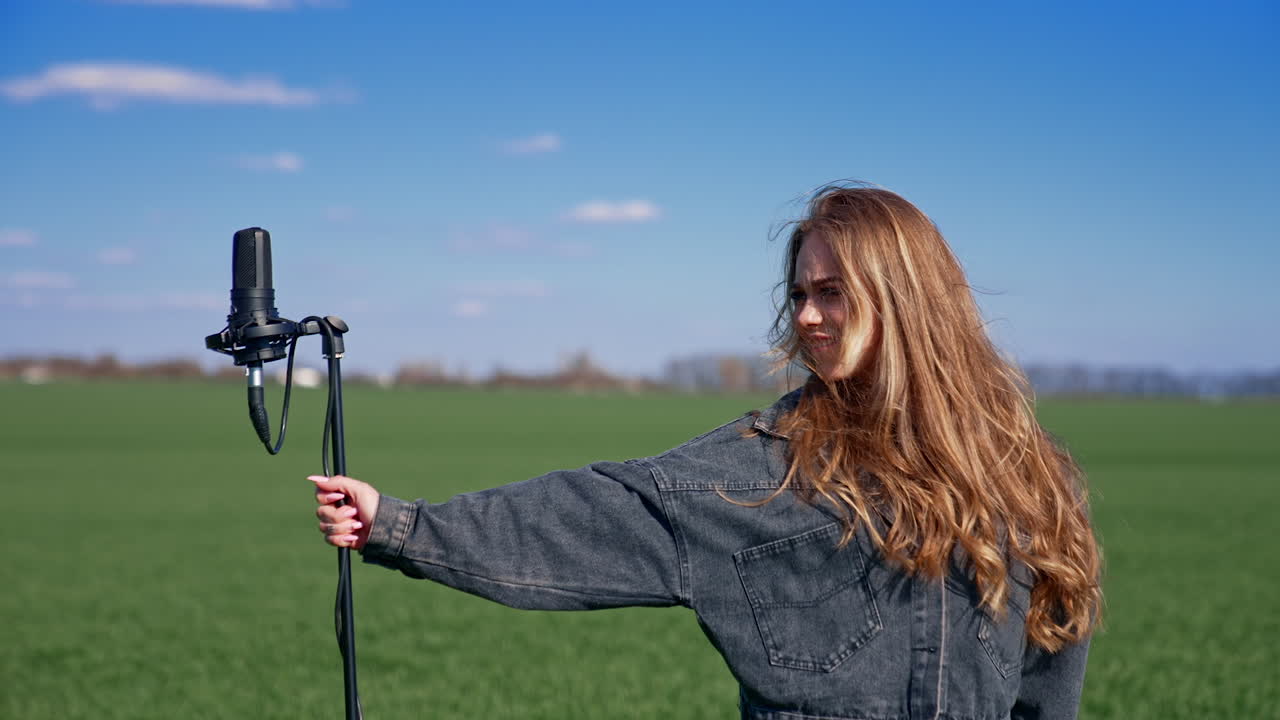 Beautiful girl near microphone outdoors. Young female singer in a field dancing and singing into microphone looking at the camera.