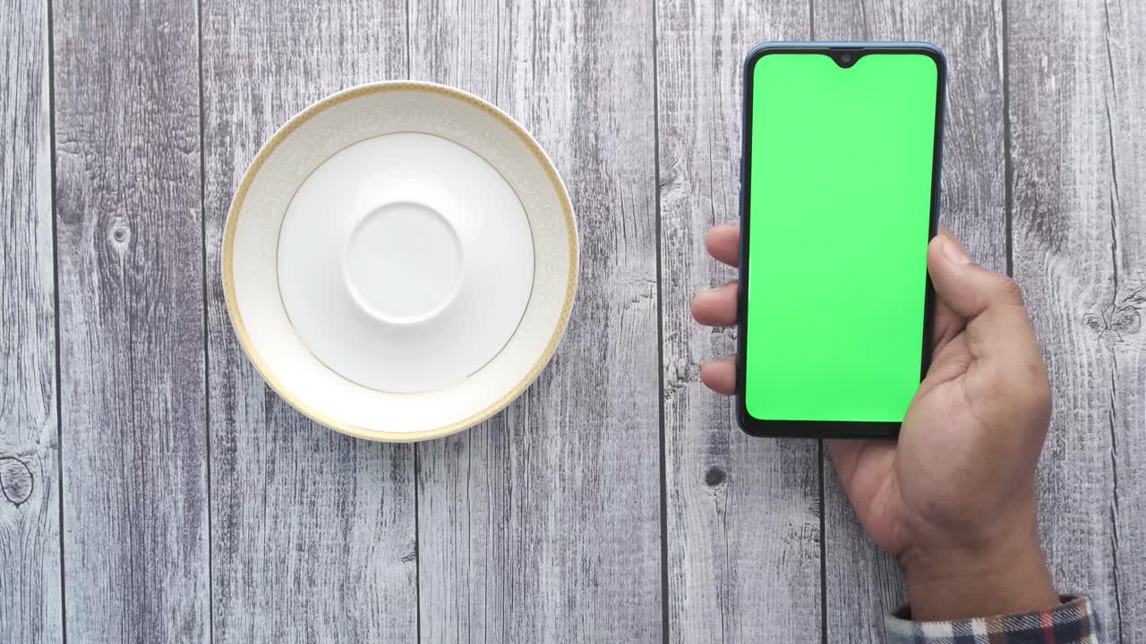 나무 테이블 위에 차 컵 에 초록색 화면을 가진 스마트폰을 들고 있는 사람