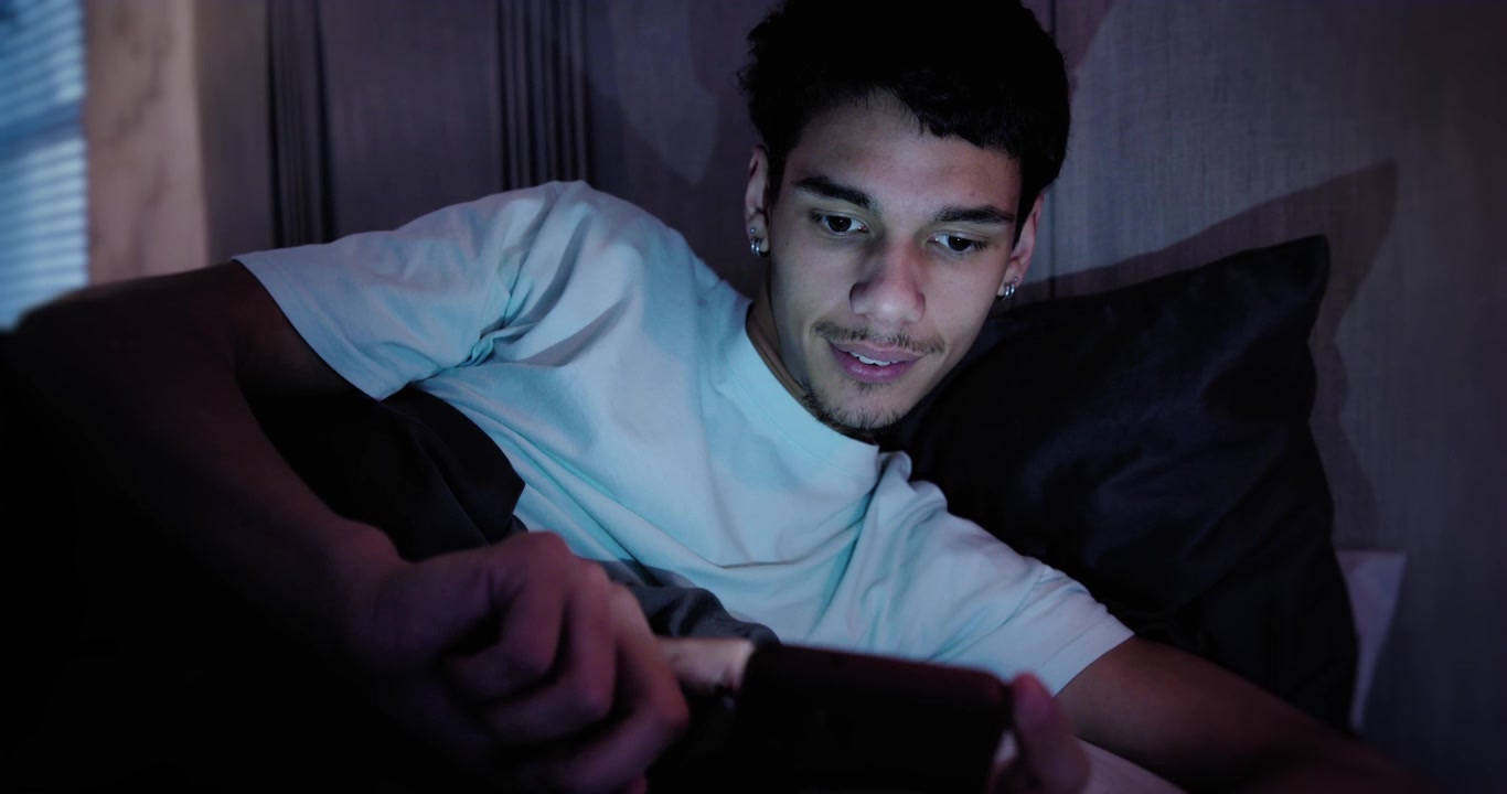 African American man lying on bed with dark pillow, phone screen drawing glance, checking messages