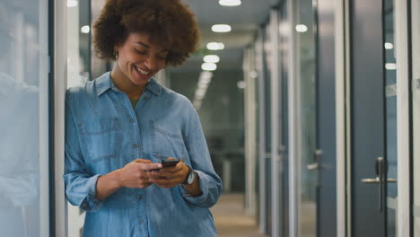 smiling businesswoman standing in corridor of modern office texting with mobile phone