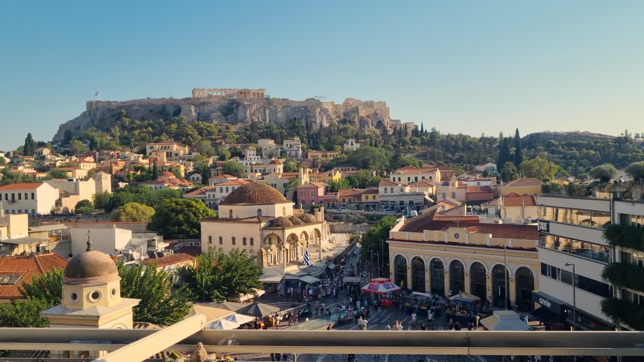 Campo lungo dell'Acropoli e del Partenone che sovrasta la trafficata Piazza Monastiraki ad Atene, con la Moschea di Tzistarakis e la metropolitana di Monastiraki sottostante, catturando la vita urbana quotidiana tra la storia antica