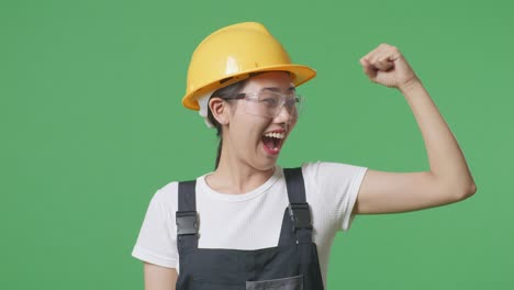 close up of asian woman worker wearing goggles and safety helmet smiling and flexing her bicep while standing in the green screen background studio