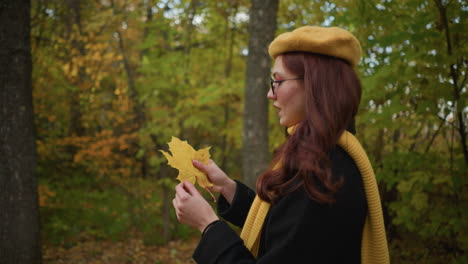 side view of lady carrying bag with warm smile holding autumn leaf, observing its details as she walks through forest surrounded by vibrant golden foliage, enjoying peaceful nature