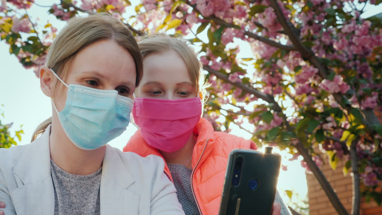 보호 마스크를 쓴 엄마와 딸이 벚꽃을 배경으로 사진을 찍습니다.