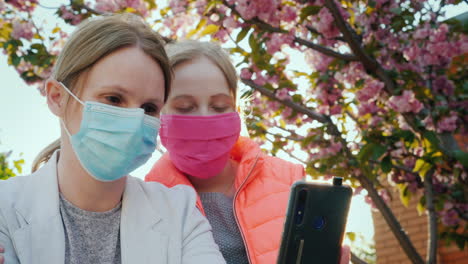 Mutter-Und-Tochter-In-Schutzmasken-Fotografieren-Sich-Vor-Dem-Hintergrund-Von-Kirschblüten