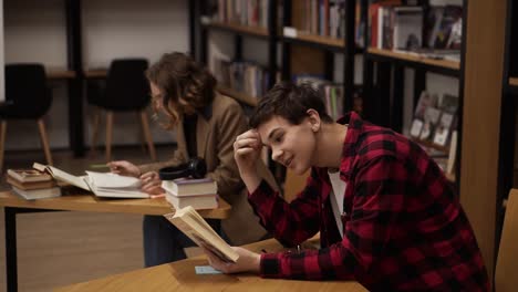 Concentrated-european-male-student-preparing-for-examination-and-reading-books-while-sitting-at-table-at-university-library-with-his-classmates-studying-books