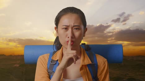 close up of asian female hiker with mountaineering backpack looking at camera and making shh gesture while standing on the top of mountain during sunset time