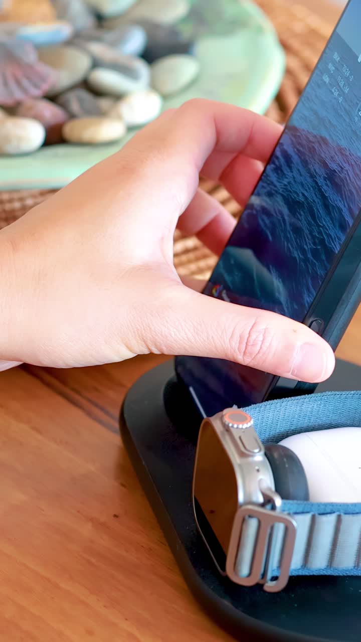 Una mano coloca un smartphone moderno en una base de carga junto a un smartwatch en un interior de hogar bien iluminado con luz natural y una superficie de madera.