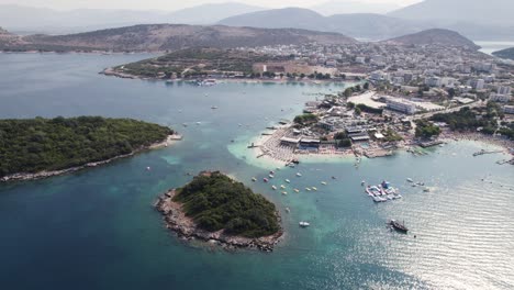 Vista-Aérea-En-órbita-De-Aguas-Serenas,-Barcos,-Islas-Exuberantes-Y-Vistas-Imponentes-De-La-Montaña-Junto-Al-Encantador-Pueblo-Ksamil-En-La-Costa-Del-Mar-Jónico