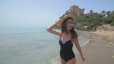 a young woman in a bikini and hat smiles as she walks along a beach with a castle in the background