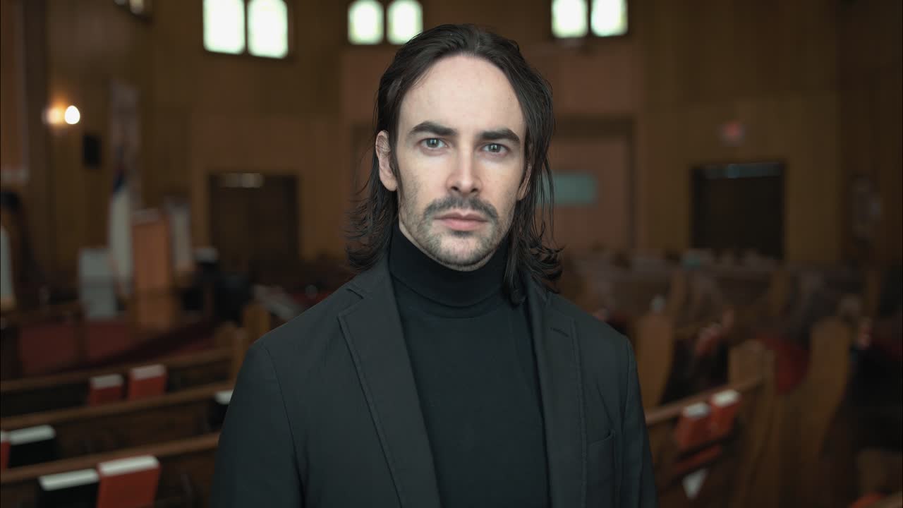 A Christian man, priest, preacher, pastor smiles in a black turtleneck and blazer stands in a church sanctuary, framed by wooden pews and soft light, creating a cinematic and contemplative mood